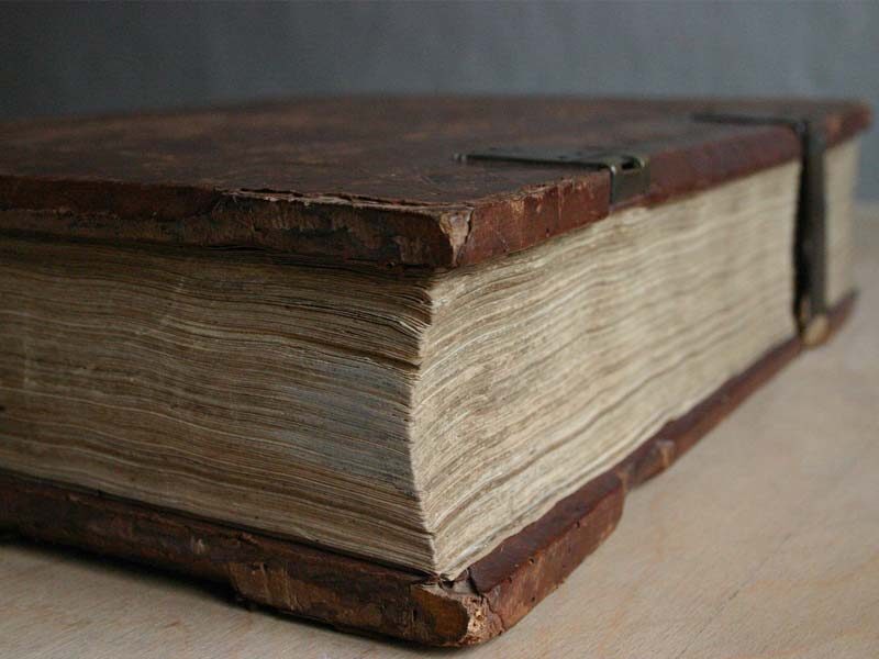 Ein dickes, altes Buch mit abgegriffenem Ledereinband und vergilbten Seiten liegt geschlossen auf einem Tisch.